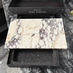 K&ouml;nnte beinhalten: Ein rechteckiges Marmortablett mit wei&szlig;en, cremefarbenen und tiefvioletten Adern. Das Tablett wird mit seiner schwarzen Verpackungsschachtel pr&auml;sentiert, auf deren Deckelseite der Text "MARBLE TRAY 20 X 30 CM" in Wei&szlig; aufgedruckt ist.