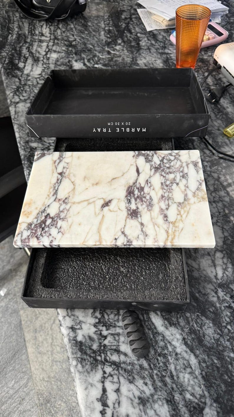 K&ouml;nnte beinhalten: Ein rechteckiges Marmortablett mit wei&szlig;en, cremefarbenen und tiefvioletten Adern. Das Tablett wird mit seiner schwarzen Verpackungsschachtel pr&auml;sentiert, auf deren Deckelseite der Text "MARBLE TRAY 20 X 30 CM" in Wei&szlig; aufgedruckt ist.