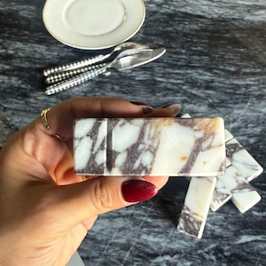 Puede incluir: Un reposacuchillos rectangular de mármol con superficie pulida, base blanca y vetas de color púrpura oscuro. El bloque de piedra tiene un acabado liso y una ranura distintiva en la parte superior para sostener la hoja del cuchillo firmemente.