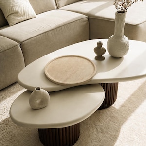 Puede incluir: Una mesa de centro de dos niveles con superficies de formas org&aacute;nicas en color blanco roto y bases cil&iacute;ndricas de color marr&oacute;n oscuro con dise&ntilde;o estriado vertical. Sobre la mesa hay una bandeja redonda beige, un jarr&oacute;n marr&oacute;n, uno blanco y uno circular.