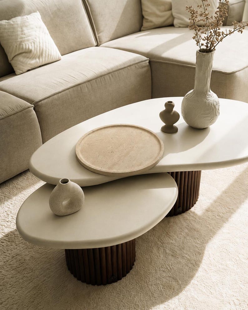 Puede incluir: Una mesa de centro de dos niveles con superficies de formas org&aacute;nicas en color blanco roto y bases cil&iacute;ndricas de color marr&oacute;n oscuro con dise&ntilde;o estriado vertical. Sobre la mesa hay una bandeja redonda beige, un jarr&oacute;n marr&oacute;n, uno blanco y uno circular.