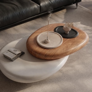 Puede incluir: Una mesa de centro de dos niveles con una superficie superior ovalada de madera y una inferior blanca. El nivel superior sostiene una bandeja circular beige con un incienso, una bandeja negra peque&ntilde;a con un objeto decorativo y un libro.