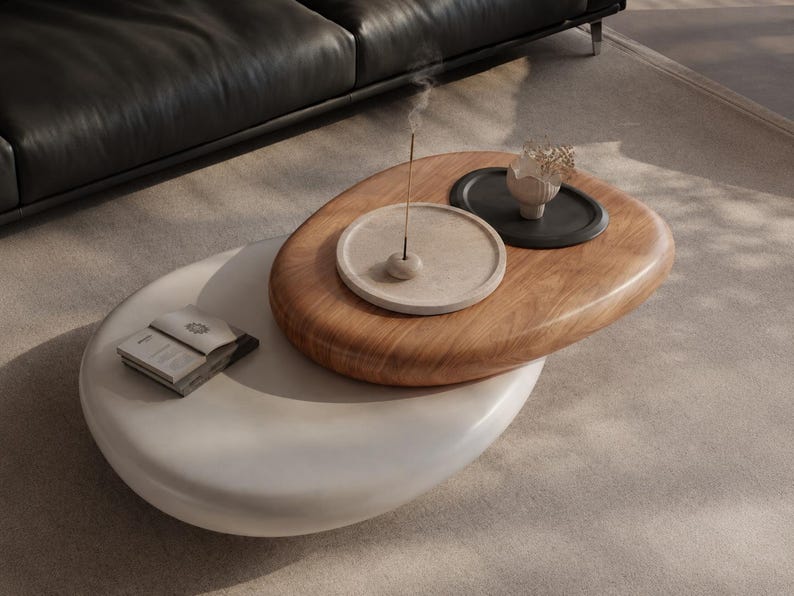 Puede incluir: Una mesa de centro de dos niveles con una superficie superior ovalada de madera y una inferior blanca. El nivel superior sostiene una bandeja circular beige con un incienso, una bandeja negra peque&ntilde;a con un objeto decorativo y un libro.