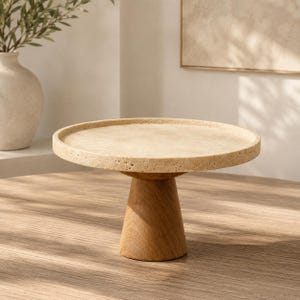Puede incluir: Un soporte circular para pasteles con una parte superior de piedra travertino beige texturizada y superficie porosa. La base está construida con madera lisa de color marrón claro y tiene una forma cónica que sostiene la plataforma de piedra plana.
