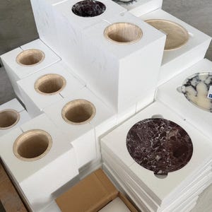 Puede incluir: Una colecci&oacute;n de recipientes cil&iacute;ndricos y platos circulares de piedra empaquetados en bloques de espuma blanca. Los art&iacute;culos de piedra presentan varios colores, incluyendo patrones de m&aacute;rmol rojo oscuro, negro, blanco y beige.