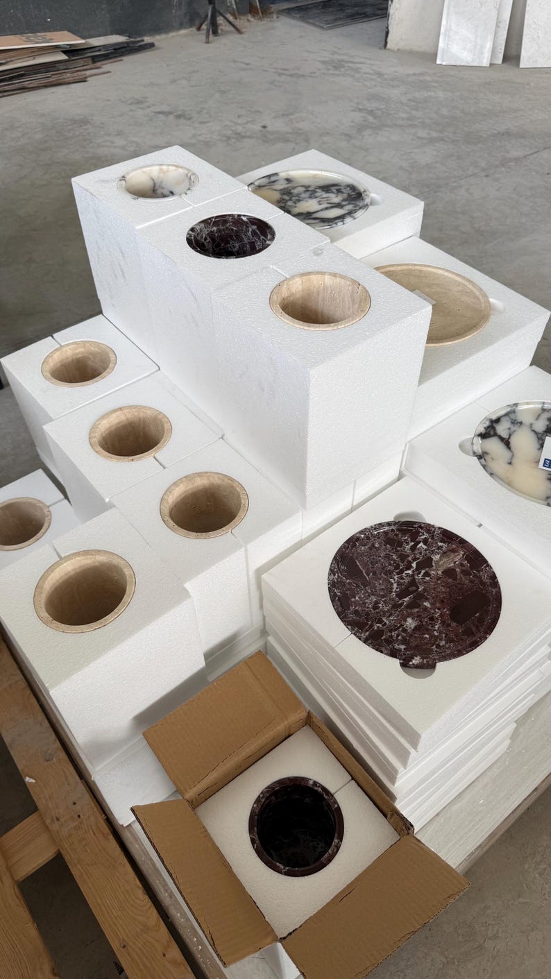 Puede incluir: Una colecci&oacute;n de recipientes cil&iacute;ndricos y platos circulares de piedra empaquetados en bloques de espuma blanca. Los art&iacute;culos de piedra presentan varios colores, incluyendo patrones de m&aacute;rmol rojo oscuro, negro, blanco y beige.