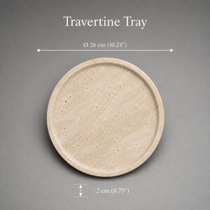 Puede incluir: Una bandeja redonda de travertino beige con una superficie porosa y texturizada. El texto Travertine Tray es visible sobre el objeto. Una l&iacute;nea de dimensi&oacute;n indica un di&aacute;metro de 26 cm y una altura de 2 cm, o 10,24 y 0,79 pulgadas.