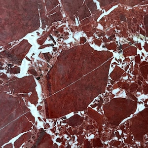 Puede incluir: Una losa de mármol rojo pulido con un patrón brechoide de fragmentos de piedra angulares en tonos carmesí y granate, unidos por una red de vetas cristalinas de color blanco brillante que crean una textura geométrica de alto contraste.