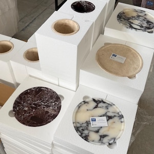 Puede incluir: M&uacute;ltiples lavabos circulares de piedra y discos planos a juego est&aacute;n dispuestos en embalajes de espuma blanca. Las piezas de piedra presentan varios patrones naturales en tonos blanco, negro, crema y granate, algunas con peque&ntilde;as etiquetas.