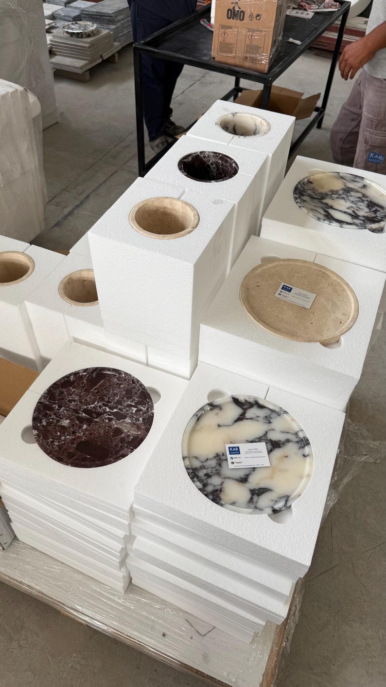 Puede incluir: M&uacute;ltiples lavabos circulares de piedra y discos planos a juego est&aacute;n dispuestos en embalajes de espuma blanca. Las piezas de piedra presentan varios patrones naturales en tonos blanco, negro, crema y granate, algunas con peque&ntilde;as etiquetas.