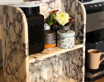 May include: A two-tiered marble shelf with a cream and brown veined pattern. The top shelf holds a black square canister with a chevron design, a small metal tin, and a round tin labelled Immuni-tea. The bottom shelf holds three clear glass mugs.