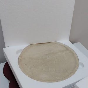 Puede incluir: Un disco circular de piedra o cer&aacute;mica de color beige claro se encuentra dentro de un inserto de embalaje protector cuadrado de espuma de poliestireno blanca. El disco tiene una superficie lisa y plana, parcialmente oculta por la espuma.