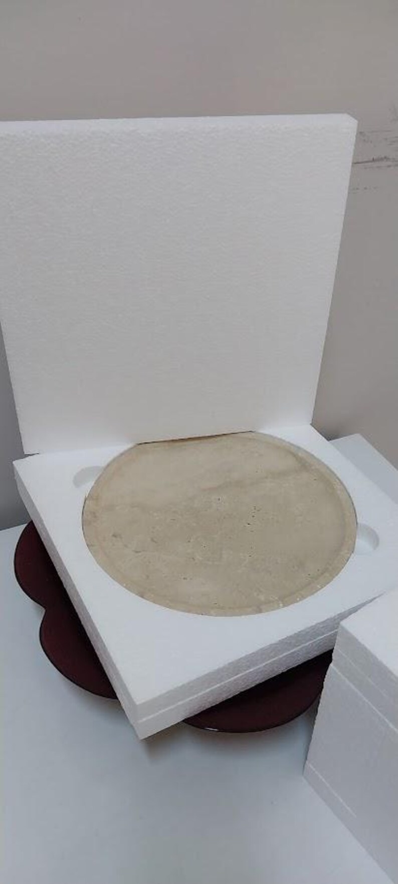 Puede incluir: Un disco circular de piedra o cer&aacute;mica de color beige claro se encuentra dentro de un inserto de embalaje protector cuadrado de espuma de poliestireno blanca. El disco tiene una superficie lisa y plana, parcialmente oculta por la espuma.