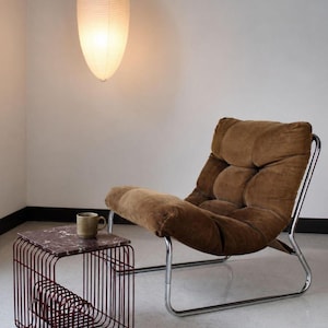 May include: A brown corduroy lounge chair with a chrome metal frame, positioned next to a small side table featuring a dark red marble top and a wire-frame base. A ceramic mug sits on the table, and a white paper pendant light hangs above.