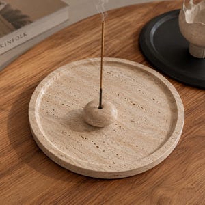 Puede incluir: Una bandeja circular para incienso hecha de travertino beige con una textura de piedra natural porosa. Una peque&ntilde;a cuenta de piedra a juego con un orificio central se sit&uacute;a en el centro de la bandeja, sosteniendo una varilla de incienso fina.