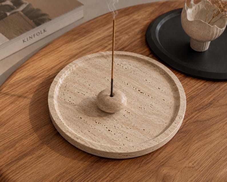 Puede incluir: Una bandeja circular para incienso hecha de travertino beige con una textura de piedra natural porosa. Una peque&ntilde;a cuenta de piedra a juego con un orificio central se sit&uacute;a en el centro de la bandeja, sosteniendo una varilla de incienso fina.