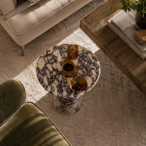 May include: A round side table featuring a white and dark purple marble pattern with a cylindrical base. On the tabletop sits a mustard yellow ceramic mug filled with a dark liquid and a small gold-toned scalloped bowl containing dark chocolate pieces.