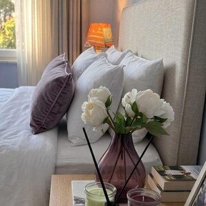 K&ouml;nnte beinhalten: Ein Schlafzimmer mit beigem Polsterkopfteil, wei&szlig;er Bettw&auml;sche und einem violetten Samtkissen. Auf einem Marmortablett stehen eine violette Glasvase mit wei&szlig;en Pfingstrosen, ein Duftdiffusor sowie zwei Kerzengl&auml;ser in Hellgr&uuml;n und Dunkelrot.