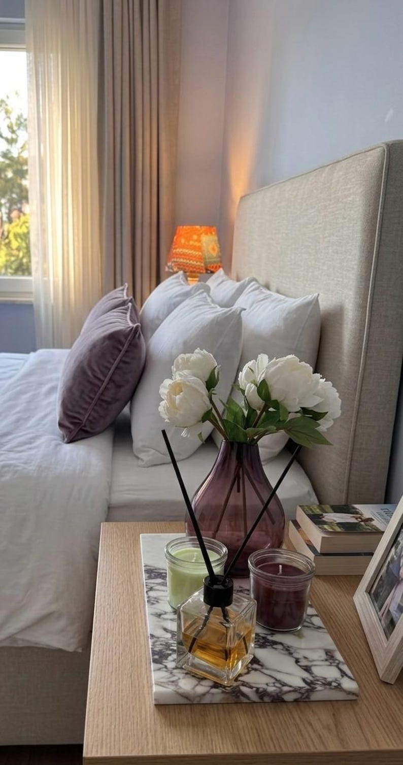K&ouml;nnte beinhalten: Ein Schlafzimmer mit beigem Polsterkopfteil, wei&szlig;er Bettw&auml;sche und einem violetten Samtkissen. Auf einem Marmortablett stehen eine violette Glasvase mit wei&szlig;en Pfingstrosen, ein Duftdiffusor sowie zwei Kerzengl&auml;ser in Hellgr&uuml;n und Dunkelrot.