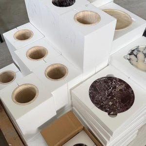 Puede incluir: Una colecci&oacute;n de recipientes cil&iacute;ndricos de piedra y platos circulares de piedra empaquetados en bloques de espuma protectora blanca. Algunos recipientes son de color marr&oacute;n oscuro o beige, mientras que los platos presentan patrones de m&aacute;rmol blanco, negro y rojo oscuro.