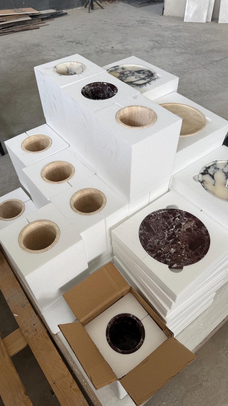 Puede incluir: Una colecci&oacute;n de recipientes cil&iacute;ndricos de piedra y platos circulares de piedra empaquetados en bloques de espuma protectora blanca. Algunos recipientes son de color marr&oacute;n oscuro o beige, mientras que los platos presentan patrones de m&aacute;rmol blanco, negro y rojo oscuro.