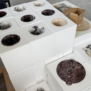 Puede incluir: Una colecci&oacute;n de bloques de espuma cuadrados blancos, cada uno con un recorte circular que contiene una pieza de piedra pulida. Las piezas muestran varios patrones de m&aacute;rmol en tonos negro, blanco y burdeos, con una abertura circular de madera.
