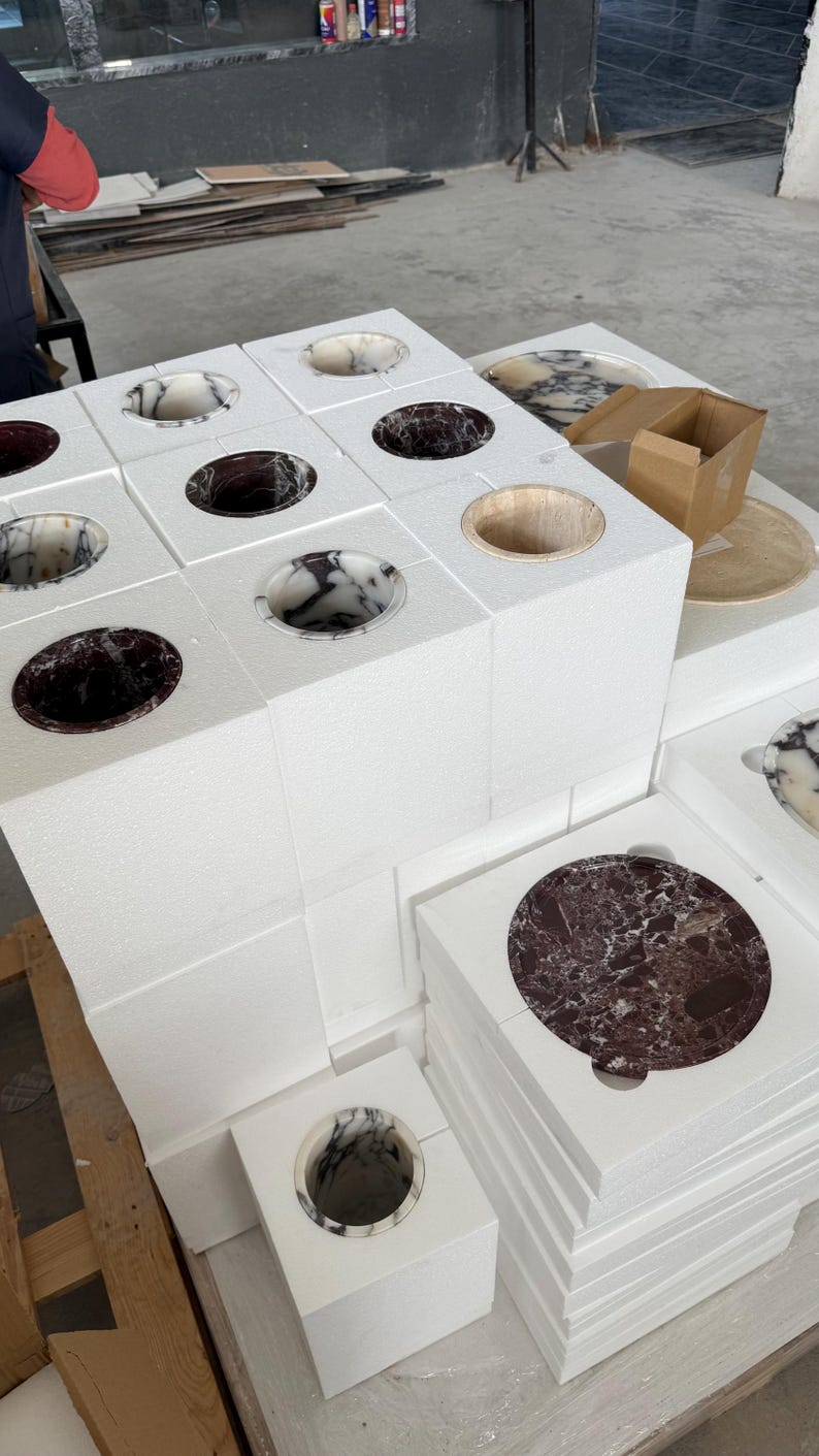 Puede incluir: Una colecci&oacute;n de bloques de espuma cuadrados blancos, cada uno con un recorte circular que contiene una pieza de piedra pulida. Las piezas muestran varios patrones de m&aacute;rmol en tonos negro, blanco y burdeos, con una abertura circular de madera.