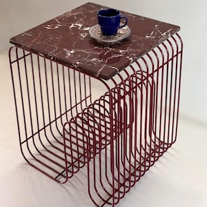 Pode incluir: Uma mesa lateral quadrada com um tampo de mármore vermelho escuro com veios brancos, apoiada por uma base construída a partir de uma série de hastes metálicas paralelas e curvas de cor vermelho escuro. Uma chávena azul está num pires.