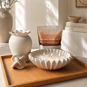 May include: A wooden tray displays a white, scalloped-edge candle bowl with three lit wicks, a decorative knot, a vase, and a bag of "Pourable Candle Sand". The arrangement is on a light surface, creating a calm, neutral-toned display.