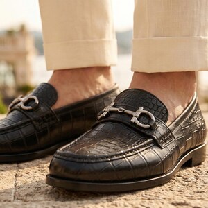 Puede incluir: Mocasines de cuero negro con textura de cocodrilo. Los zapatos tienen una hebilla plateada y se usan con pantalones color crema. La imagen está tomada al aire libre sobre una superficie de piedra.