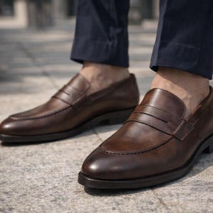 May include: Brown leather penny loafers with a classic design. The shoes have a rounded toe, a strap across the vamp, and a low heel. The loafers are worn with navy blue trousers.