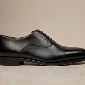 May include: A single black leather Oxford shoe with a cap toe. The shoe has a classic design with a lace-up closure and a low heel. The shoe is approximately 30 cm long.
