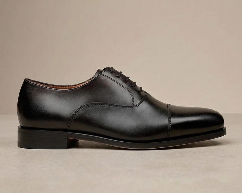 May include: A single black leather Oxford shoe with a cap toe. The shoe has a classic design with a lace-up closure and a low heel. The shoe is approximately 30 cm long.