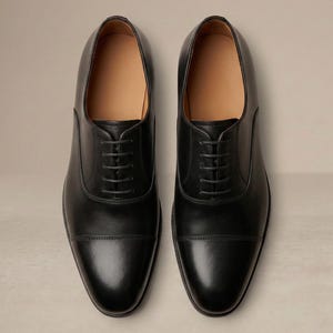 May include: A pair of black leather Oxford shoes with a cap-toe design and lace-up fastening. The shoes have a polished finish and a tan interior. The image is a top-down view, highlighting the symmetrical design. The shoes are a classic style.