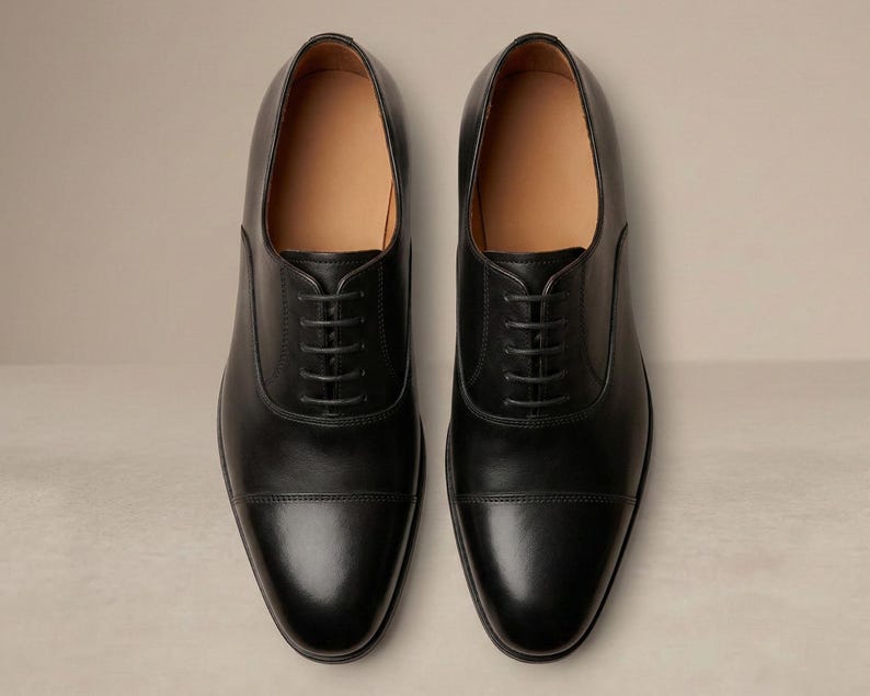 May include: A pair of black leather Oxford shoes with a cap-toe design and lace-up fastening. The shoes have a polished finish and a tan interior. The image is a top-down view, highlighting the symmetrical design. The shoes are a classic style.