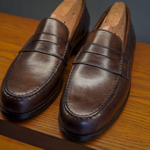 May include: Two brown leather penny loafers with wooden shoe trees. The shoes are presented on a wooden surface, highlighting their polished appearance and traditional design. The stitching and craftsmanship are clearly visible. The shoes are approximately 30 cm long.