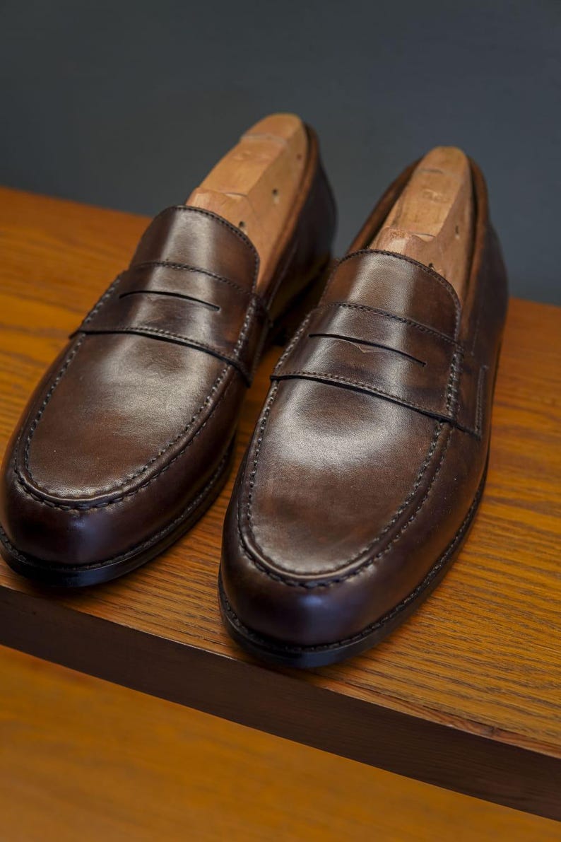 May include: Two brown leather penny loafers with wooden shoe trees. The shoes are presented on a wooden surface, highlighting their polished appearance and traditional design. The stitching and craftsmanship are clearly visible. The shoes are approximately 30 cm long.