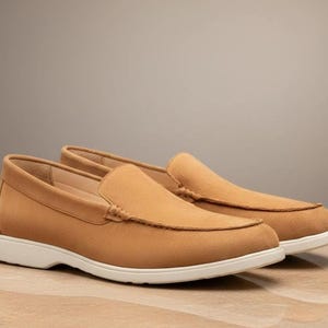 May include: A pair of tan suede loafers with a classic design. The shoes have a rounded toe, a slip-on style, and a white sole. The loafers are displayed on a light-colored surface.
