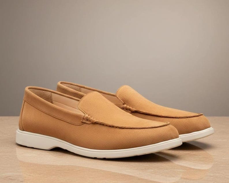 May include: A pair of tan suede loafers with a classic design. The shoes have a rounded toe, a slip-on style, and a white sole. The loafers are displayed on a light-colored surface.