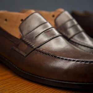 May include: Close-up of two brown leather penny loafers. The shoes have a classic design with a strap across the vamp and visible stitching. The leather has a polished finish. Shoe trees are visible in the background.