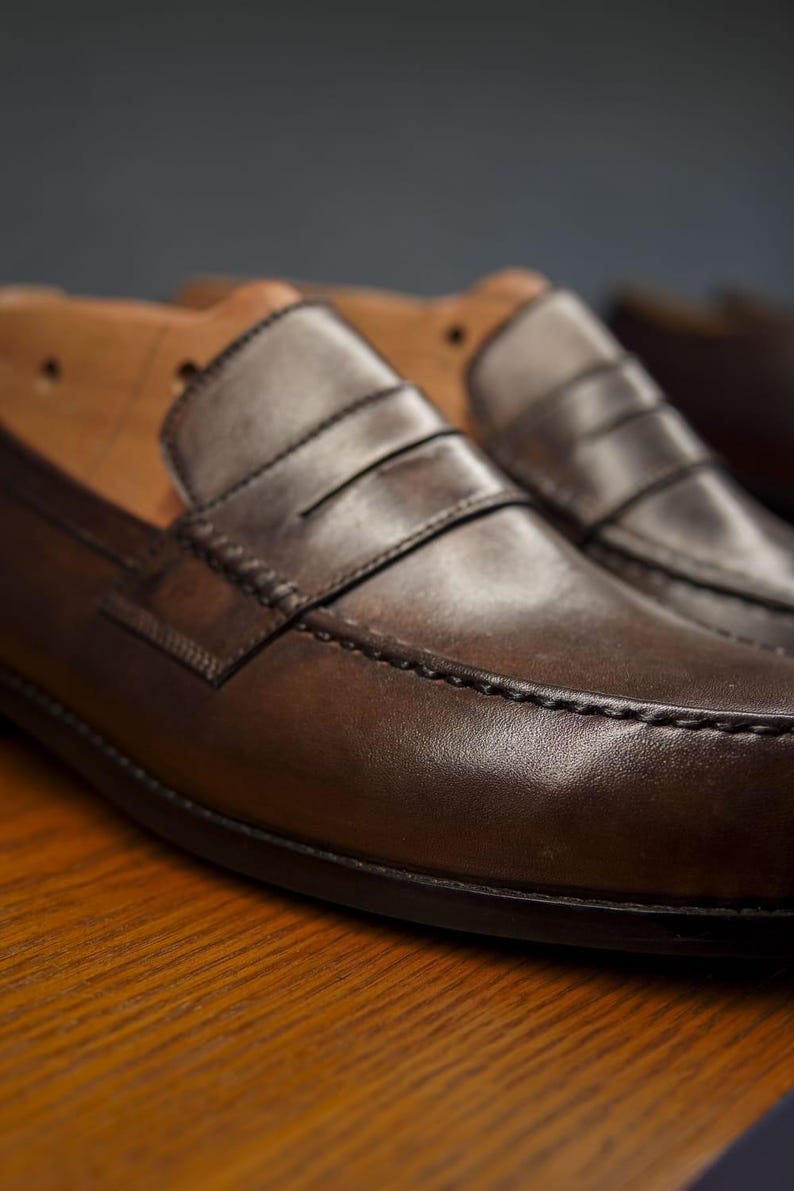 May include: Close-up of two brown leather penny loafers. The shoes have a classic design with a strap across the vamp and visible stitching. The leather has a polished finish. Shoe trees are visible in the background.