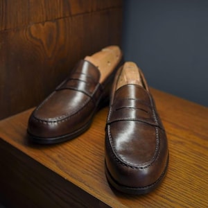 May include: A pair of brown leather penny loafers with wooden shoe trees. The shoes are displayed on a wooden surface, highlighting their polished finish and classic design. The loafers have a rounded toe and a strap across the vamp.