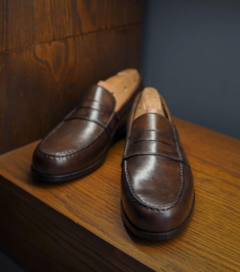 May include: A pair of brown leather penny loafers with wooden shoe trees. The shoes are displayed on a wooden surface, highlighting their polished finish and classic design. The loafers have a rounded toe and a strap across the vamp.