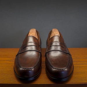 May include: A pair of brown leather penny loafers with wooden shoe trees inside. The shoes are displayed on a wooden surface against a dark grey background. The loafers have a classic design with a strap across the vamp.