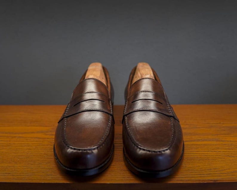May include: A pair of brown leather penny loafers with wooden shoe trees inside. The shoes are displayed on a wooden surface against a dark grey background. The loafers have a classic design with a strap across the vamp.