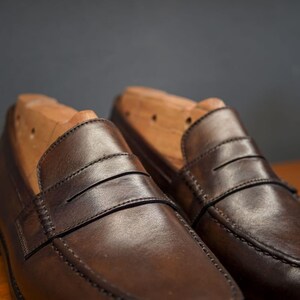 May include: A pair of brown leather penny loafers with wooden shoe trees. The shoes feature a classic design with a strap across the vamp and visible stitching. The leather has a polished finish.