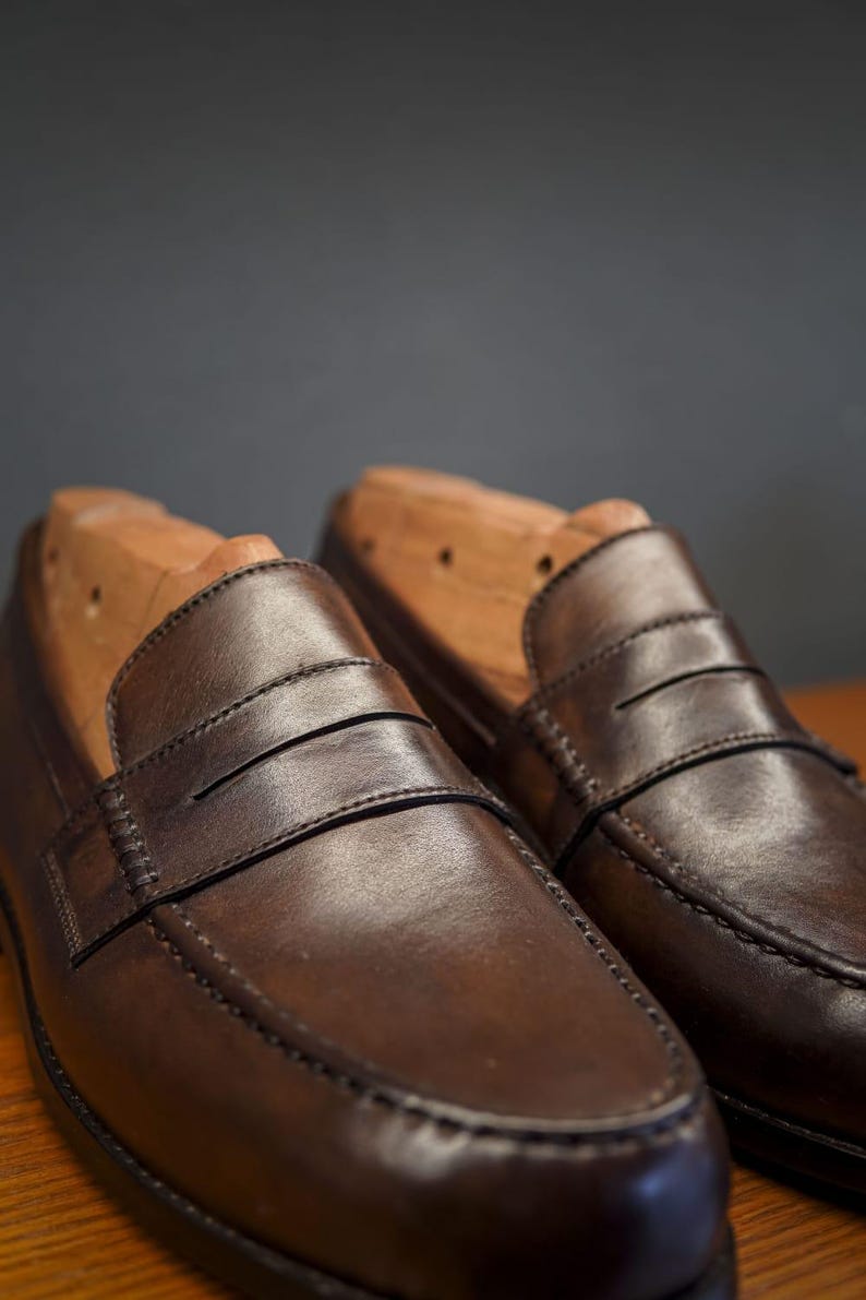 May include: A pair of brown leather penny loafers with wooden shoe trees. The shoes feature a classic design with a strap across the vamp and visible stitching. The leather has a polished finish.