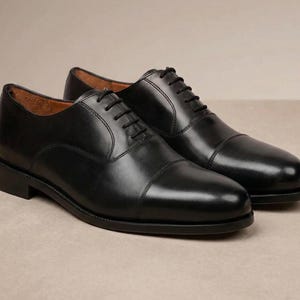May include: A pair of black leather Oxford shoes. The shoes have a cap toe design and black laces. The shoes are displayed on a neutral background. The shoes are a classic style.