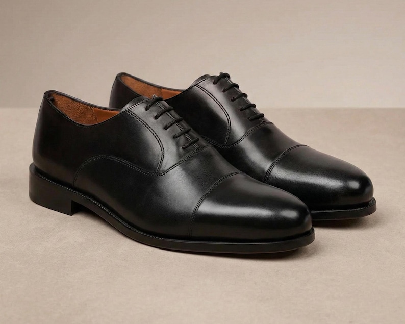 May include: A pair of black leather Oxford shoes. The shoes have a cap toe design and black laces. The shoes are displayed on a neutral background. The shoes are a classic style.