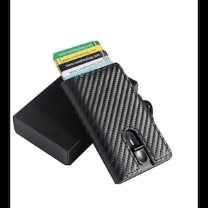 Può includere: Un portafoglio nero in fibra di carbonio con un porta carte, accanto a una scatola nera. Sono visibili diverse carte, con l'indirizzo del sito web www.casekeybag.com stampato sopra. Il portafoglio ha una chiusura a bottone.